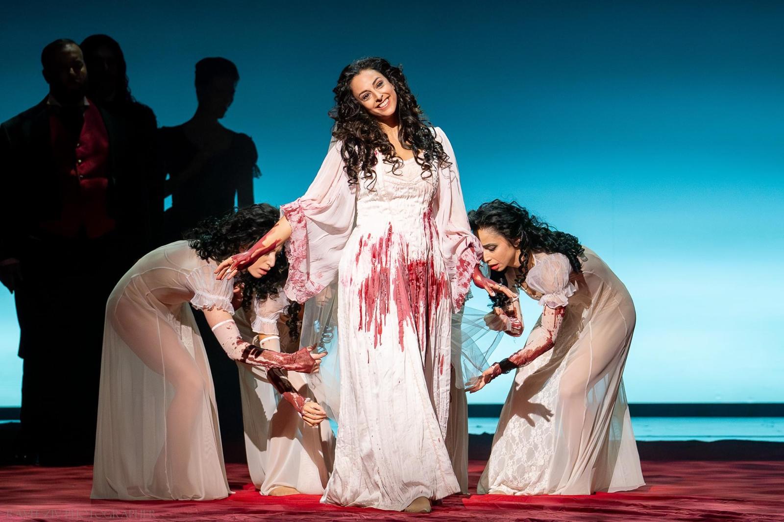 Hila Fahima in the title role of Lucia di Lammermoor, Tel Aviv 2024 — standing between two dancers in a blood-stained gown, photo by Ofer Rosner