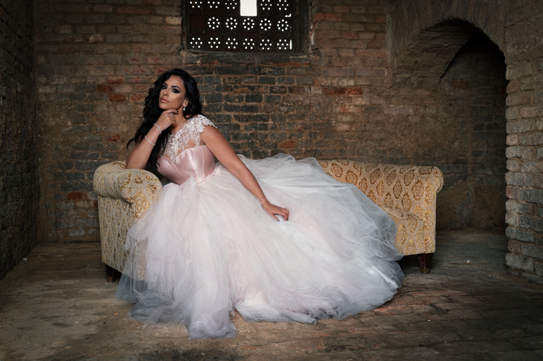 Hila Fahima in a blush tulle ball gown on a brocade sofa in a vaulted brick room, by Manfred Baumann, 2025