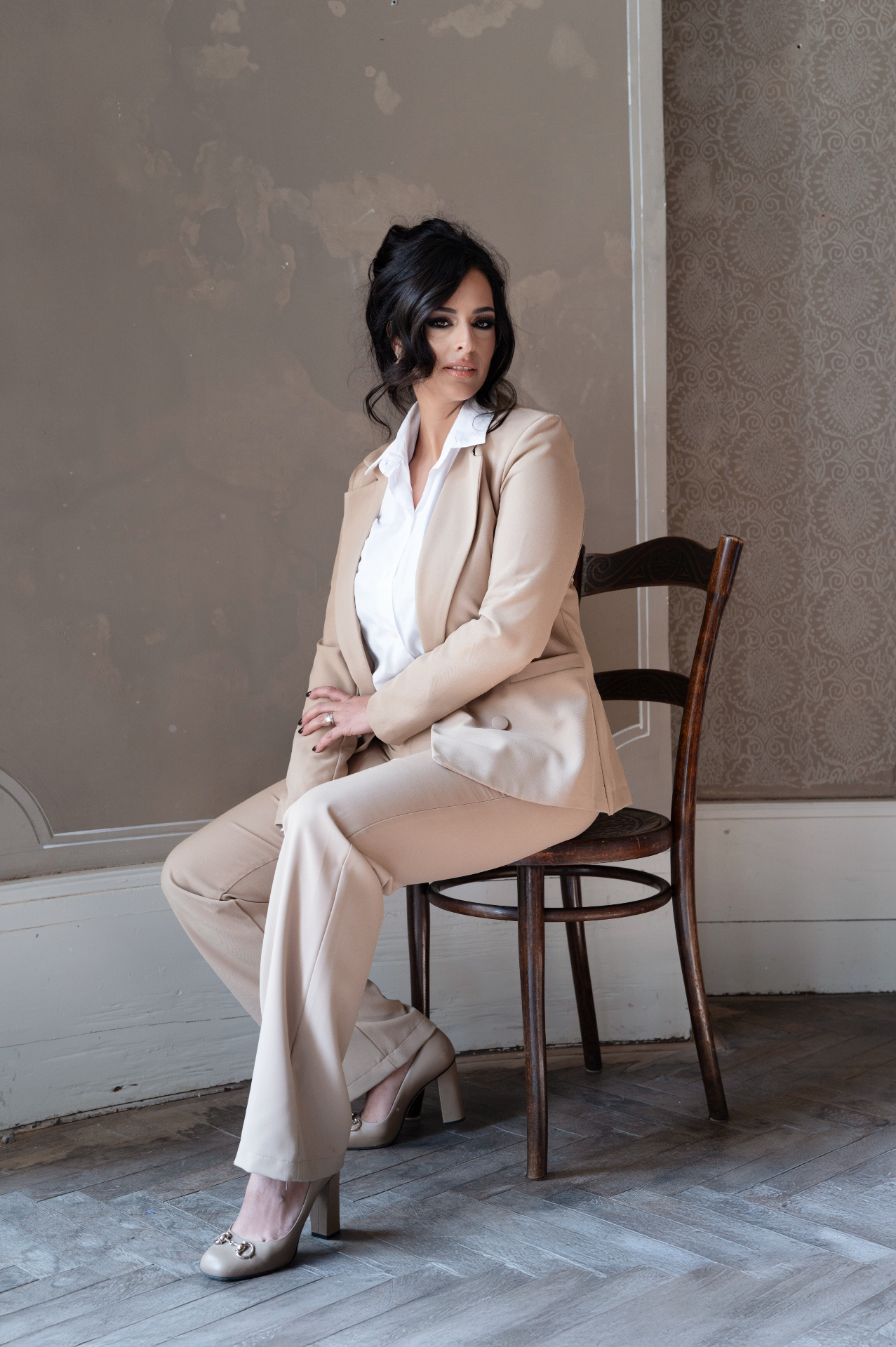 Hila Fahima seated on a bentwood chair in a beige suit and white shirt, by Manfred Baumann, 2025
