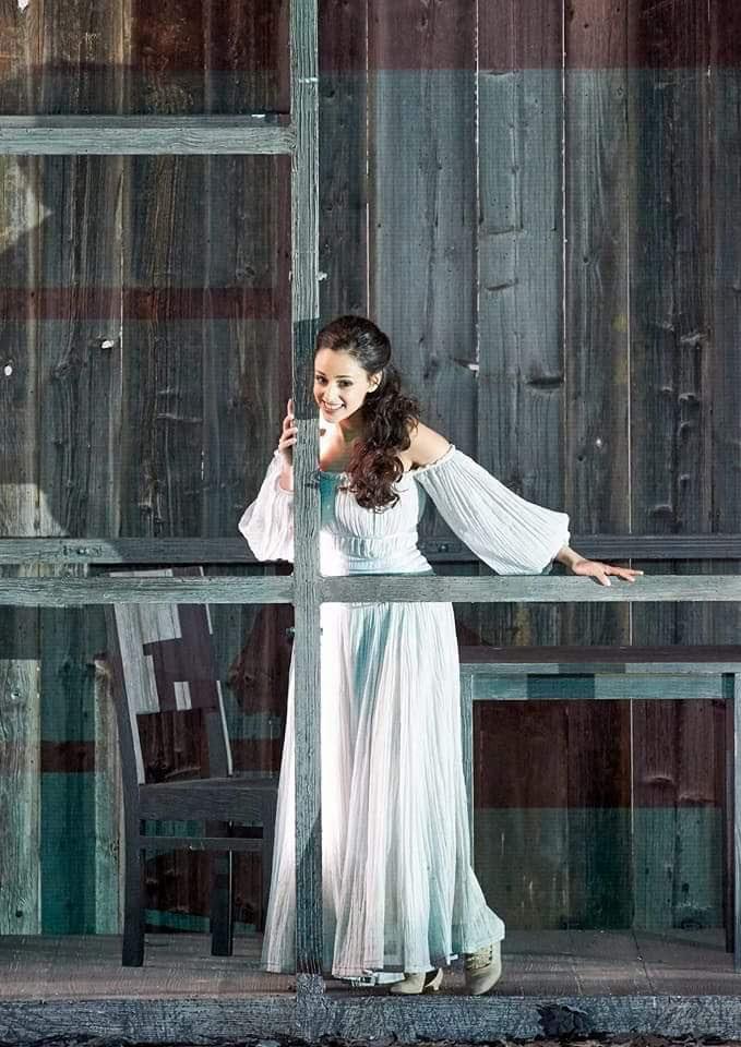 Hila Fahima as Gilda in Rigoletto at the Vienna State Opera — in a white gown leaning against a wooden beam, photo by Michael Pöhn
